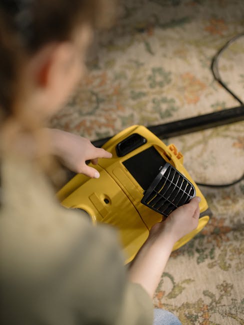 A person kneeling on a decorative patterned carpet in a room, using a yellow and black vacuum cleaner for surface cleaning or deep cleaning. The individual, with curly hair and wearing a beige jacket 