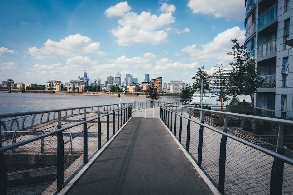 A panoramic view of a modern urban cityscape featuring a calm river flanked by tall, contemporary office and residential buildings constructed from glass, steel, and concrete. The buildings vary in he