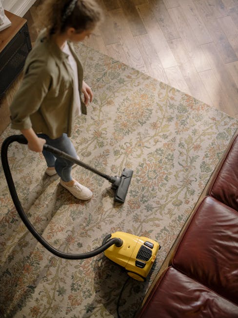 A person using a yellow vacuum cleaner on a traditional patterned area rug in a living room, with light wood flooring and a red leather sofa visible. The individual is wearing casual clothing, including a tan jacket, and is performing domestic surface cleaning to remove dust and debris. The room is well-lit, with natural light highlighting the cleanliness of the space. This image demonstrates the detailed deep cleaning services offered by Carpet Cleaners SE8, specializing in same day carpet cleaning quotes and availability in the Docklands area.