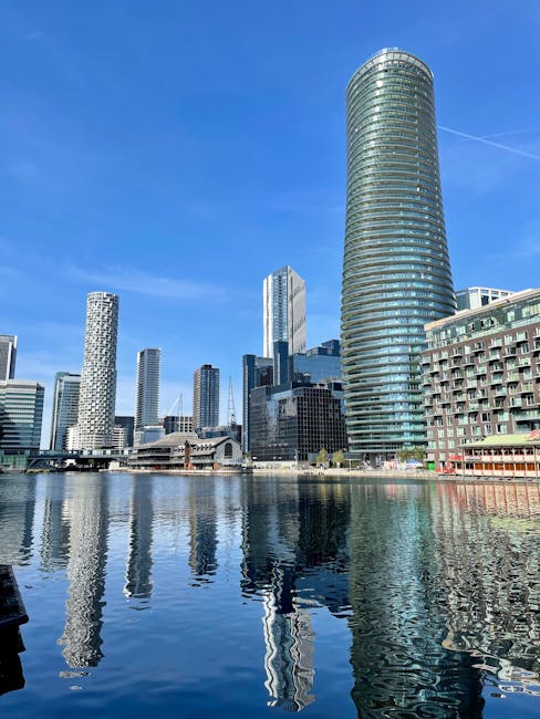 A modern cityscape featuring a variety of tall commercial and residential buildings with glass facades reflecting the blue sky and clouds, situated along a waterway with calm, mirror-like surface. The tallest building is a cylindrical skyscraper with a rounded top, surrounded by other high-rise structures of different architectural styles, including rectangular and patterned designs. The area appears clean and well-maintained, with natural daylight illuminating the scene. This urban environment exemplifies contemporary architectural design, with the water providing a picturesque foreground. Carpet Cleaners SE8 specializes in surface cleaning and deep cleaning services for both residential and commercial properties, ensuring hygiene and cleanliness in such vibrant city settings.